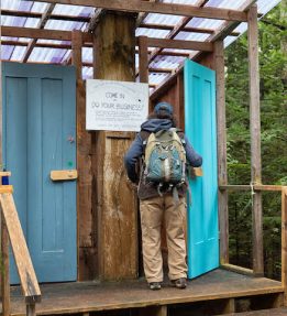 A fairgoer wearing a ballcap and a backpack opens the blue door to an outhouse