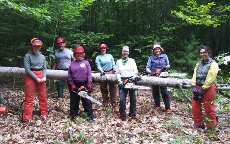 A group of people smiling while holding logs and chainsaws.